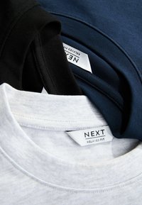 Tři trička v černé, námořnické a bílé barvě. Každé má kulatý výstřih a štítky s nápisem "NEXT RELAXED FIT." Viditelná struktura bavlny.