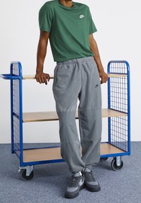 Osoba v zeleném tričku Nike, šedých fleecových kalhotách Nike a šedých teniskách Nike se opírá o modrý kovový vozík s dřevěnými policemi.