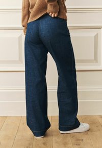 Platmalu džinsi tumši zila krāsā ar teksturētu apdari, ar kontrastējošiem diegiem un valkāti ar baltām kedām uz koka grīdas.