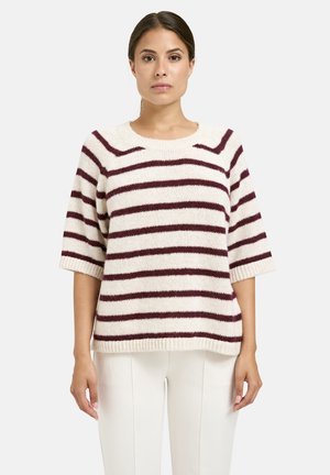 Woman wearing a cream sweater with maroon horizontal stripes and elbow-length sleeves, paired with white pants, standing against a plain background.