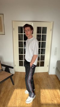 Noormees, kellel on käed taskutes, kannab valget särk, mustasid dressipükse logoga triibuga ja valgeid tenniseid, seisab siseruumis klaaspaneelidega ukse kõrval.