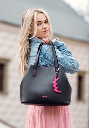 Schwarze Leder-Tote-Bag mit strukturiertem Design, rosa Reißverschlussanhänger und Logodetail. Modell trägt eine Jeansjacke und einen hellrosa Rock.