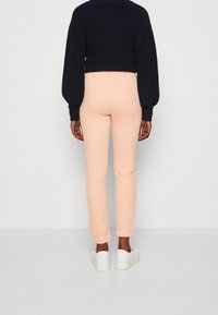 Persoon in lichtroze aansluitende joggingsbroek, zwarte cropped trui en witte sneakers, staand tegenover een witte muur.