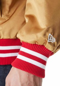 Giacca bomber in satin oro con polsini a righe rosse. I polsini presentano strisce bianche e un logo brandizzato. Texture liscia con aperture elastiche.