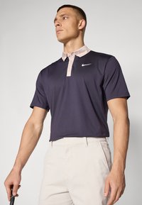 Sötétkék Nike póló, rózsaszín gallérral, rövid ujjú, fehér logóval. Bézs nadrággal párosítva, golfütő a kezében.