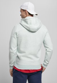 Felpa verde menta chiaro realizzata in materiale morbido, con cappuccio regolabile e polsini a coste. Indossata sopra una maglietta rossa, abbinata a pantaloni scuri.