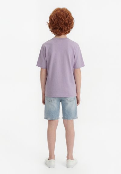 Child with curly red hair standing facing away, wearing a lavender t-shirt, light denim shorts, and white sneakers.