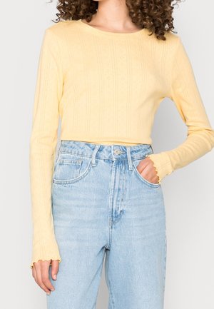 Femme portant un haut à manches longues jaune pâle avec des motifs subtils et un jean denim taille haute bleu clair, main dans la poche.