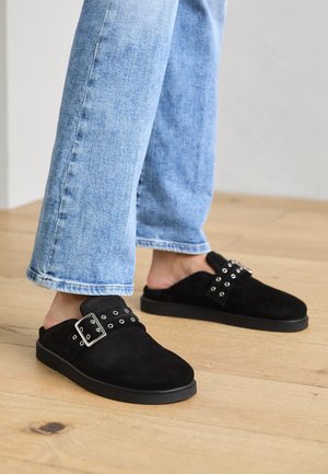 Voeten met zwarte slip-on schoenen met zilveren gesp en oogjesdetails, gecombineerd met lichtblauwe rechte spijkerbroek, staand op houten vloer.