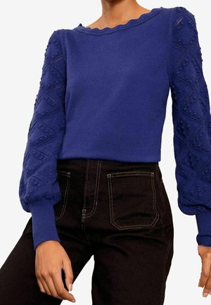 Blauer Strickpullover mit überschnittenen, strukturierten Ärmeln und einem gewellten Ausschnitt, getragen zu hoch taillierten schwarzen Hosen mit weißen Nähten.