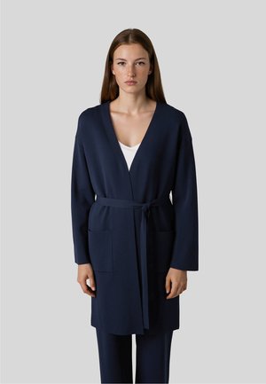 Frau mit glattem braunem Haar, die eine marineblaue Strickjacke mit Gürtel über einem weißen Oberteil und passenden marineblauen Hosen vor einem grauen Hintergrund trägt.