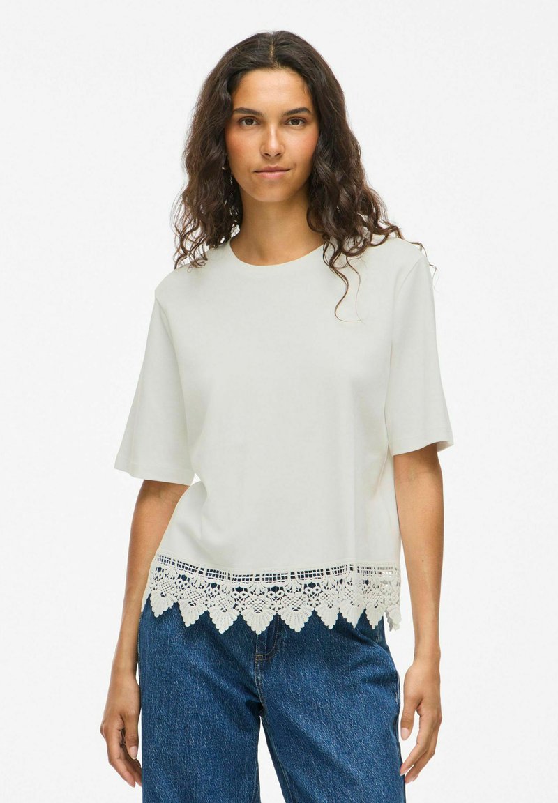 Top corto de manga corta en color beige claro con escote redondo y detalle de dobladillo de encaje con borde en forma de ondas. Combinado con jeans azules. Textura suave.