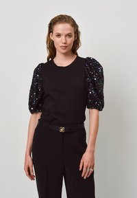 Zwarte t-shirt met textuur en pailletten pofmouwen in meerdere kleuren. Heeft een ronde halslijn en een nauwsluitend silhouet, gedragen met zwarte high-waisted broek.