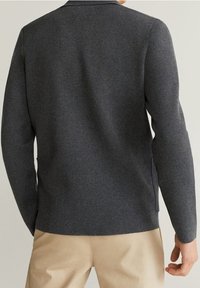 Uomo che indossa un maglione grigio scuro a maniche lunghe e pantaloni beige, visto da dietro su uno sfondo chiaro e semplice.