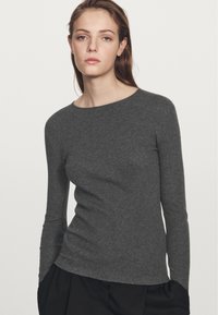 Grijze lange mouwen top in een ribbelstructuur, met een boot halslijn, slim fit en zijsplitten. Draagt met zwarte high-waisted broeken.