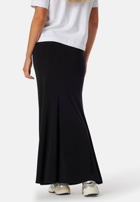 Zwarte, elegante maxi-jurk gemaakt van rekbaar materiaal, met een uitlopende onderkant en een gladde textuur. Draag een witte blouse en zilveren sneakers.