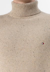 Beige garbónyakú pulóver, strukturált kötött anyagból készült, foltos mintával és egy kis logóval a bal oldalon.