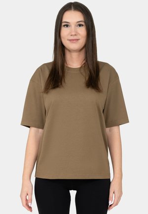 Woman standing facing forward wearing a loose brown short-sleeve shirt and black pants against a plain light background.