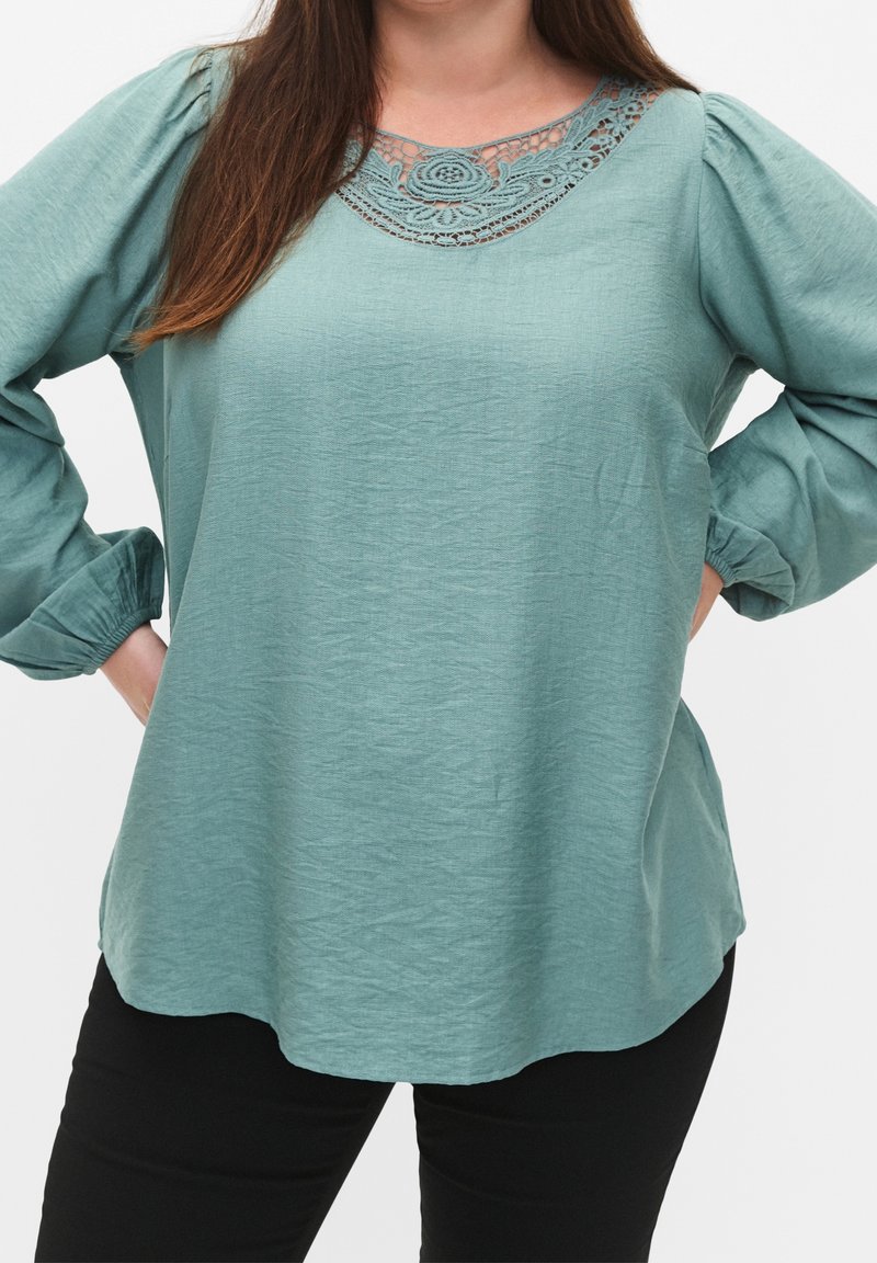 Blouse verte en tissu texturé, col rond avec des détails en dentelle intricats, et manches longues avec poignets froncés.