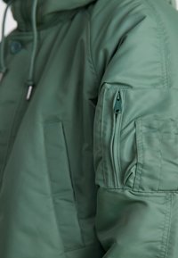 Grüne Steppjacke mit hohem Kragen, ausgestattet mit einer Reißverschlusstasche am Ärmel und Seitentaschen, aus glattem, glänzendem Stoff gefertigt.