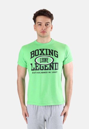 Jonge man staat recht vooruit kijkend, draagt een felgroen T-shirt met de tekst "Boxing Legend Leone 1947" en lichtgrijze joggingbroek.