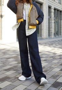 Veste matelassée à imprimé léopard avec des accents jaunes, associée à une chemise à carreaux claire et un pantalon en denim foncé à jambes larges, complétée par des baskets blanches.