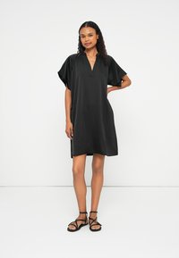 Vestido preto com mangas curtas e levemente esvoaçantes, decote em V e um corte solto, feito de um tecido suave. Usado com sandálias pretas de tiras.