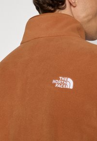 Veste en polaire marron avec un col montant et un logo blanc brodé au dos. Texture douce et design minimaliste.