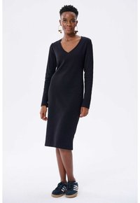 Schwarzes, geripptes Langarmkleid mit V-Ausschnitt, figurbetontem Design und knielanger Saum. Kombiniert mit dunklen Turnschuhen mit weißen Streifen.