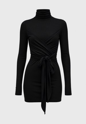Femme aux cheveux bruns et lisses portant une robe noire à manches longues, col roulé, ceinture à la taille et sandales plates noires, debout sur un fond uni.