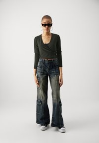 Haut long à manches ribbed vert foncé, jeans cargo bleus décolorés taille haute avec poches, et baskets Converse noires. Lunettes de soleil noires élégantes portées.
