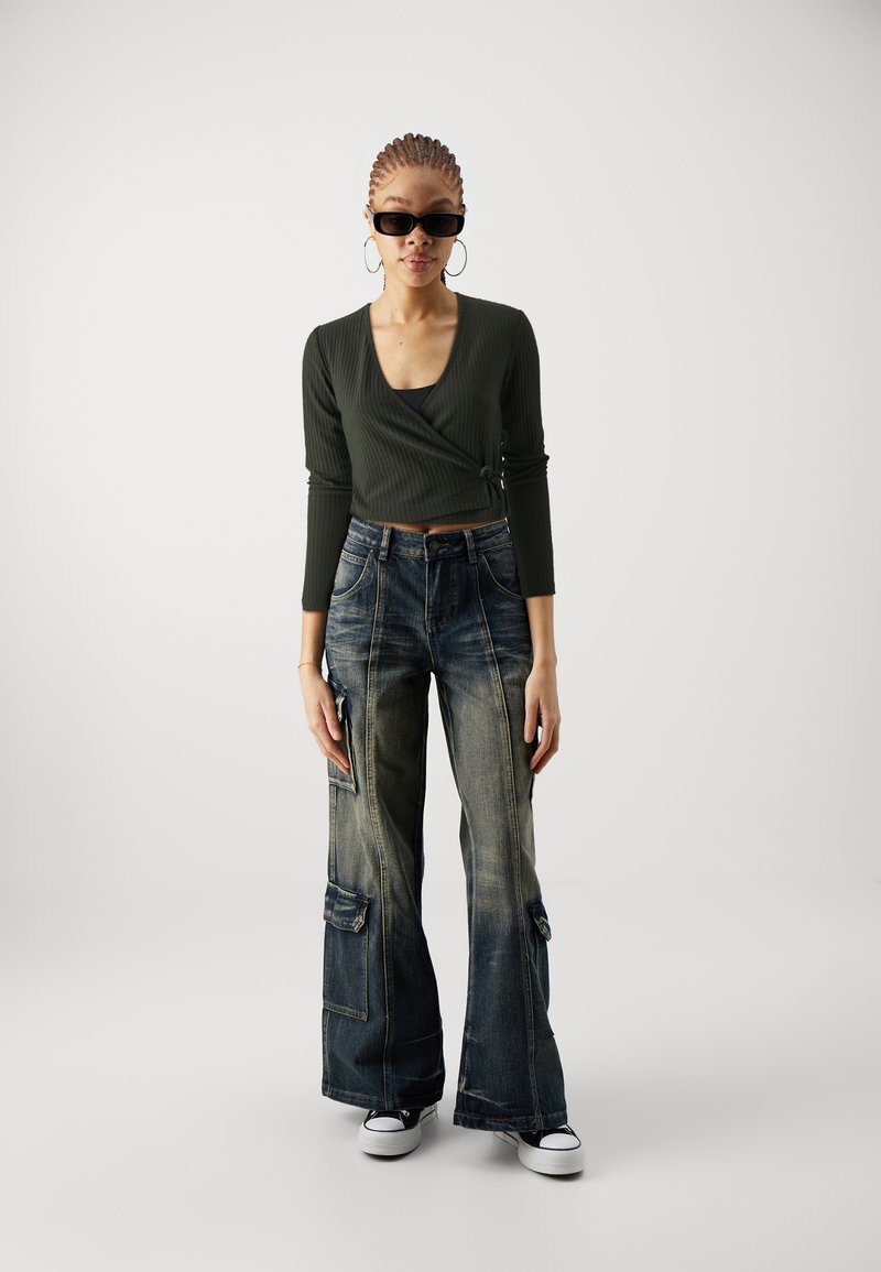 Dunkelgrünes, geripptes Langarmoberteil, hoch taillierte, ausgebleichte blaue Cargo-Jeans mit Taschen und schwarze Converse-Sneaker. Elegante schwarze Sonnenbrille getragen.