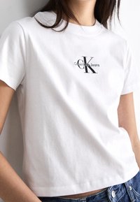 Žena v bílém tričku s krátkým rukávem s logem "Calvin Klein Jeans CK", v kombinaci s modrými džínami.