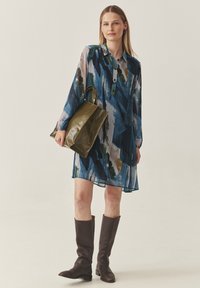 Durchsichtiges, gemustertes Hemdkleid in Blau, Grün und Beige mit einem knöpfbaren Kragen. Kombiniert mit einer dunkelolivgrünen Tragetasche und braunen knieb hohen Stiefeln.