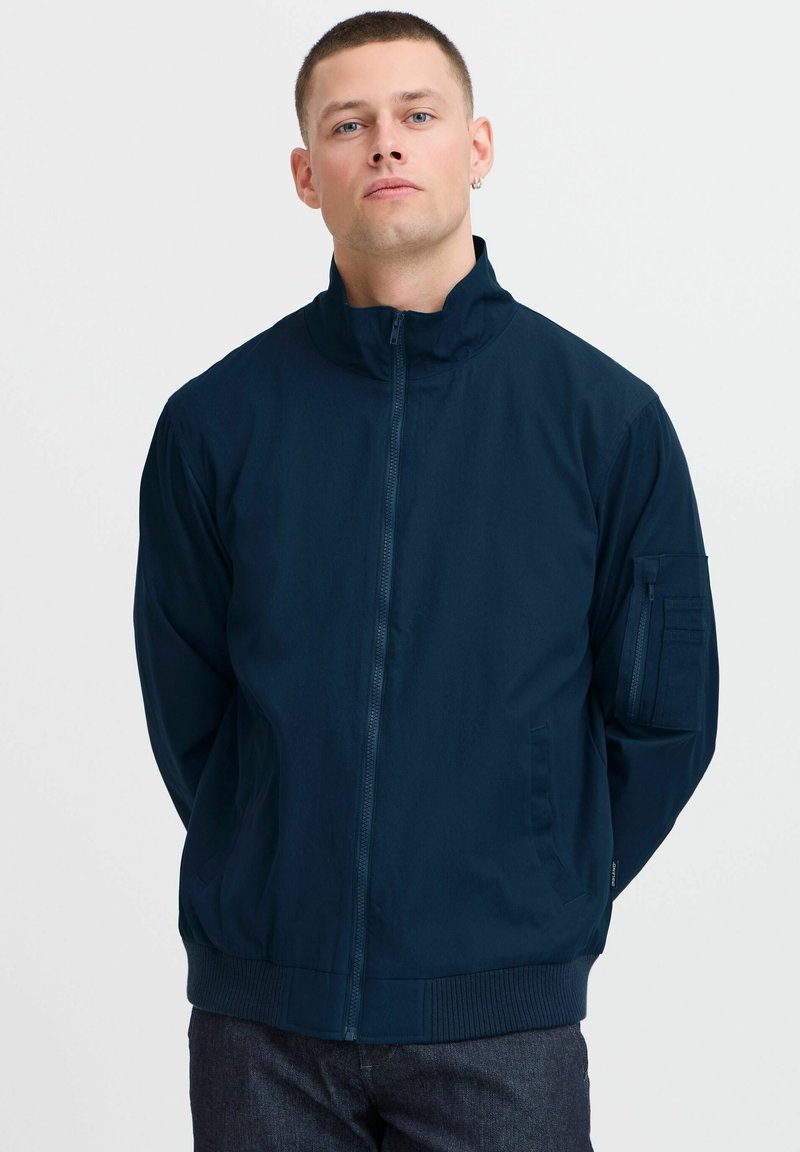 Jūrų laivyno zip-up jaketa su aukštu apykakle, glotniais rankogaliais ir apatine dalimi, turinti naudingą kišenę ant rankovės ir lygų, matinį paviršių.