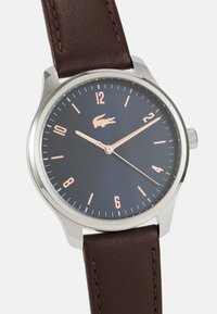 Montre à bracelet en cuir marron avec un cadran texturé bleu foncé, un boîtier en acier argenté et des index et aiguilles en or rose. Présente un logo crocodile.
