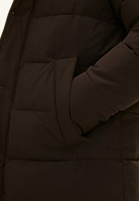 Giubbotto in piuma marrone con un'esterno liscio e lucido. Presenta una grande tasca e pannelli cuciti per l'isolamento. Maniche leggermente gonfie.