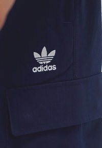 Navy blue adidas shorts with a large front pocket and a white trefoil logo, featuring three horizontal stripes and a textured fabric.