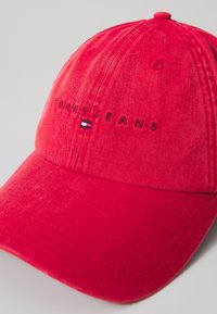 Red cotton baseball cap featuring "TOMMY JEANS" embroidered in black with a small blue, white, and red logo accent on the front.