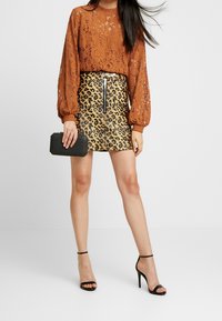 Frau trägt eine rötlich-braune schicke Bluse mit langen Ärmeln aus Spitze, einen Minirock mit Leopardenmuster und Frontreißverschluss, schwarze High-Heels und hält eine schwarze Clutch.