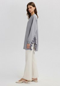 Maglia grigia chiara a maniche lunghe con dettaglio arricciato laterale, abbinata a pantaloni a gamba larga color crema e scarpe slingback di colore chiaro. Tessuto morbido.