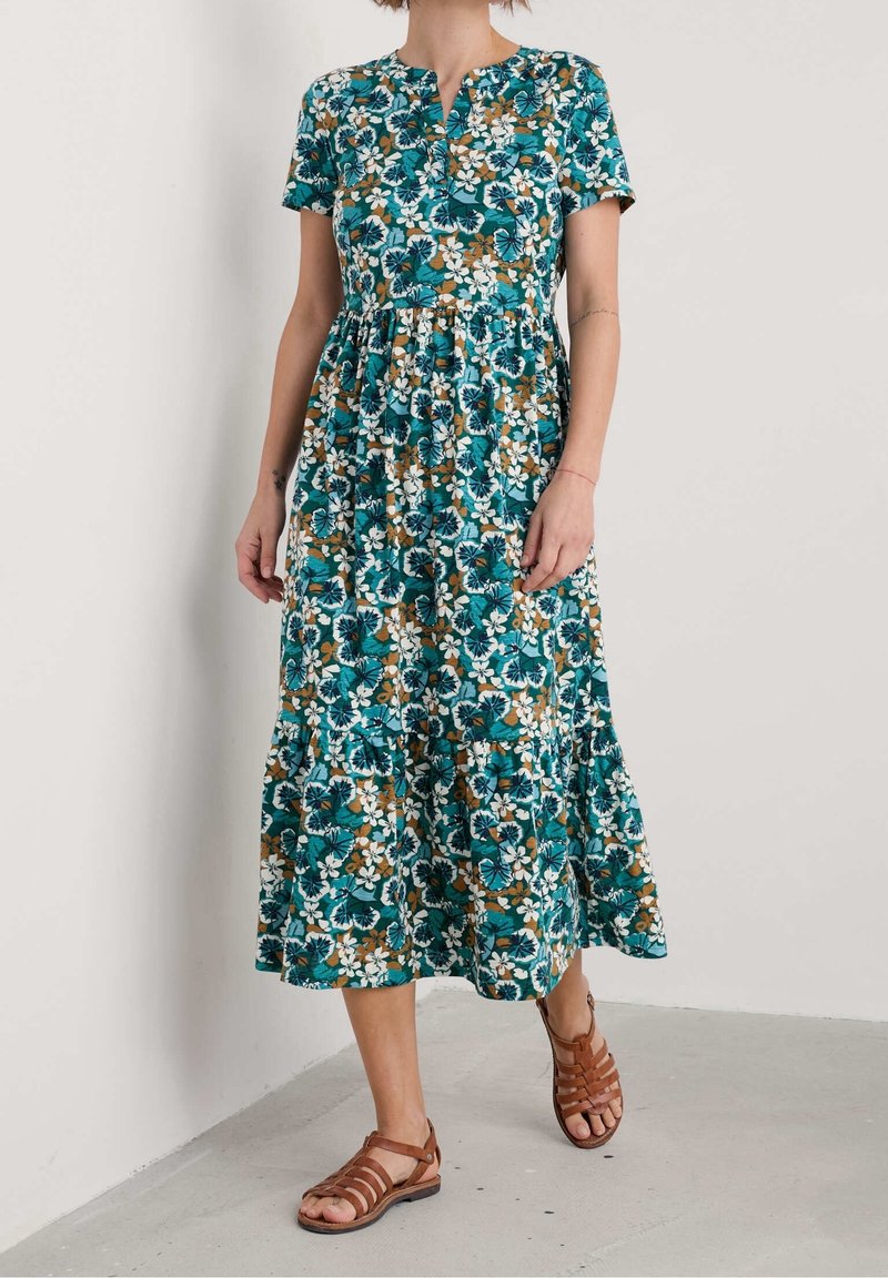 Femme portant une robe midi à manches courtes fleurie turquoise, blanche et marron avec un ourlet volanté et des sandales marron à brides.
