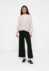 Cardigan de punto beige claro con seis botones, combinado con pantalones de pierna ancha negros. La modelo sostiene un pequeño bolso blanco texturizado, llevando zapatos y calcetines negros.