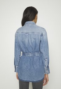 Ljusblå denimskjorta med midjebälte, långa ärmar och knäppbara manschetter. Har en klassisk krage och bakre okdesign.