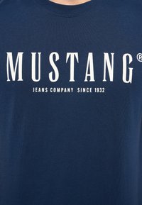 Tumši zilas kokvilnas T-krekls ar apaļu kakla izgriezumu. Uz krūšdaļas ir balts teksts "MUSTANG JEANS COMPANY SINCE 1932". Mīksta tekstūra.