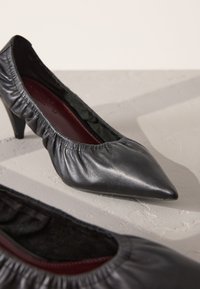 Black leather pointed-toe pump with gathered elastic around the opening, low heel, and soft maroon lining. Textured surface with sheen.