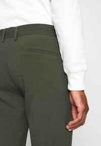 Pantaloni verde oliva con una texture liscia, dotati di una tasca posteriore e passanti per cintura, abbinati a una camicia bianca a maniche lunghe.