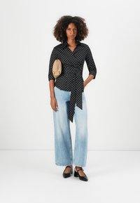 Blusa nera a pois con vita legata e maniche a tre quarti, abbinata a jeans a gamba larga di colore blu chiaro e scarpe con tacco nere.