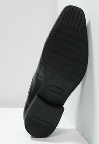 Semelle de chaussure en cuir noir avec un design texturé et décoré. Présente une surface lisse et un bloc rectangulaire au talon pour la stabilité.