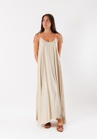 Maxi dress beige con spalline sottili, caratterizzato da una silhouette fluida e una texture liscia e morbida. Paio di sandali marroni in basso.